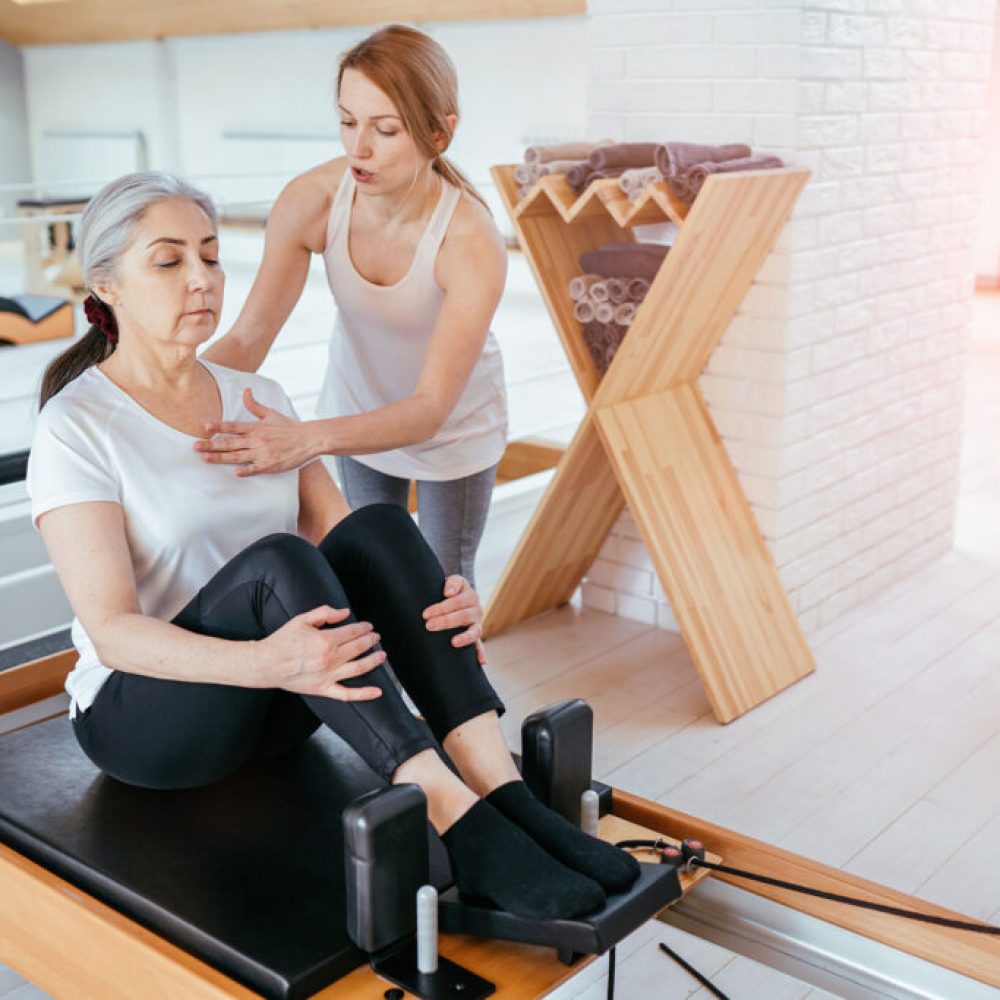 Brunette female instructor with cute two braids hairstyle consulting hansdome beard man workout pilates on reformer practice in pilates studio, working out indoor, correcting beginners, full length.