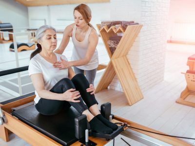 Brunette female instructor with cute two braids hairstyle consulting hansdome beard man workout pilates on reformer practice in pilates studio, working out indoor, correcting beginners, full length.
